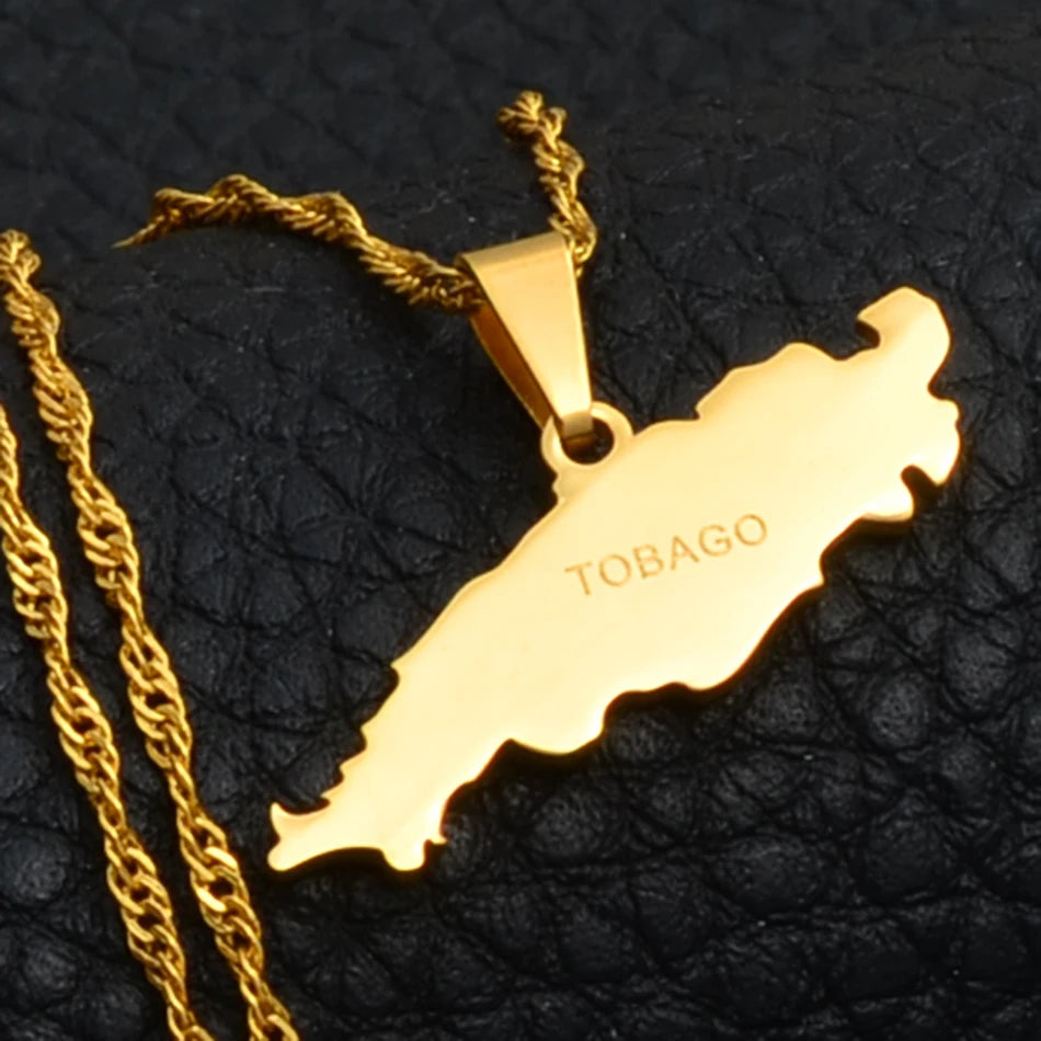 Gold map-shaped pendant necklace engraved with the word 'TOBAGO', displayed on a textured black surface next to a gold chain.