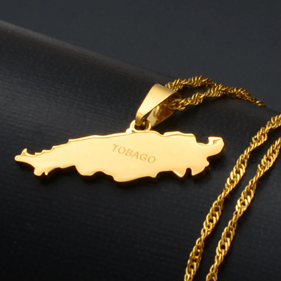 A gold-colored necklace featuring a pendant shaped like the map of Tobago, with the name engraved on the front, resting on a dark textured surface.